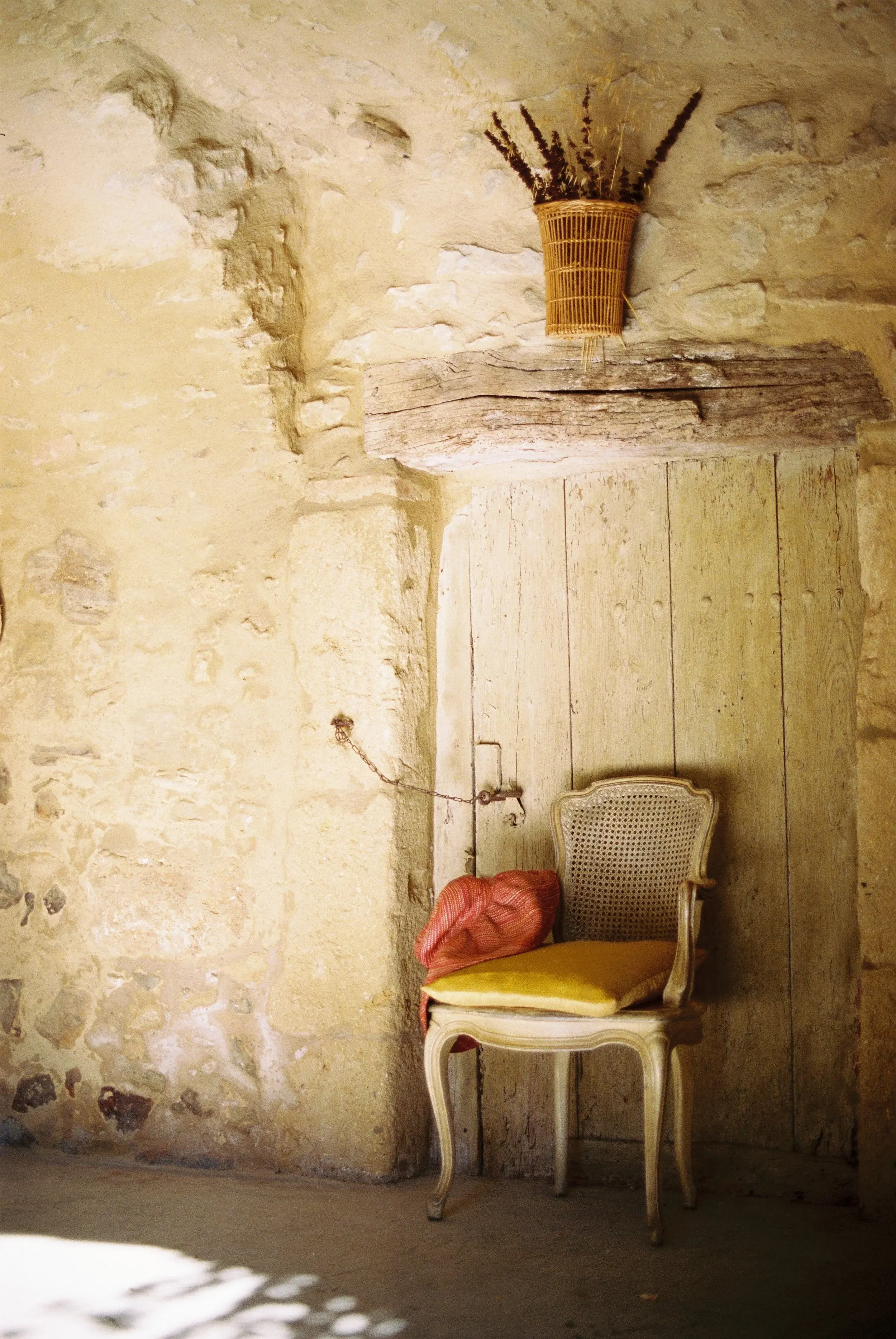 Une vieille chaise en bois avec un coussin jaune, placée contre un mur en pierre, avec un sac en tissu rouge posé dessus. Au-dessus, un panier en rotin avec des herbes séchées. La porte derrière la chaise est en bois.