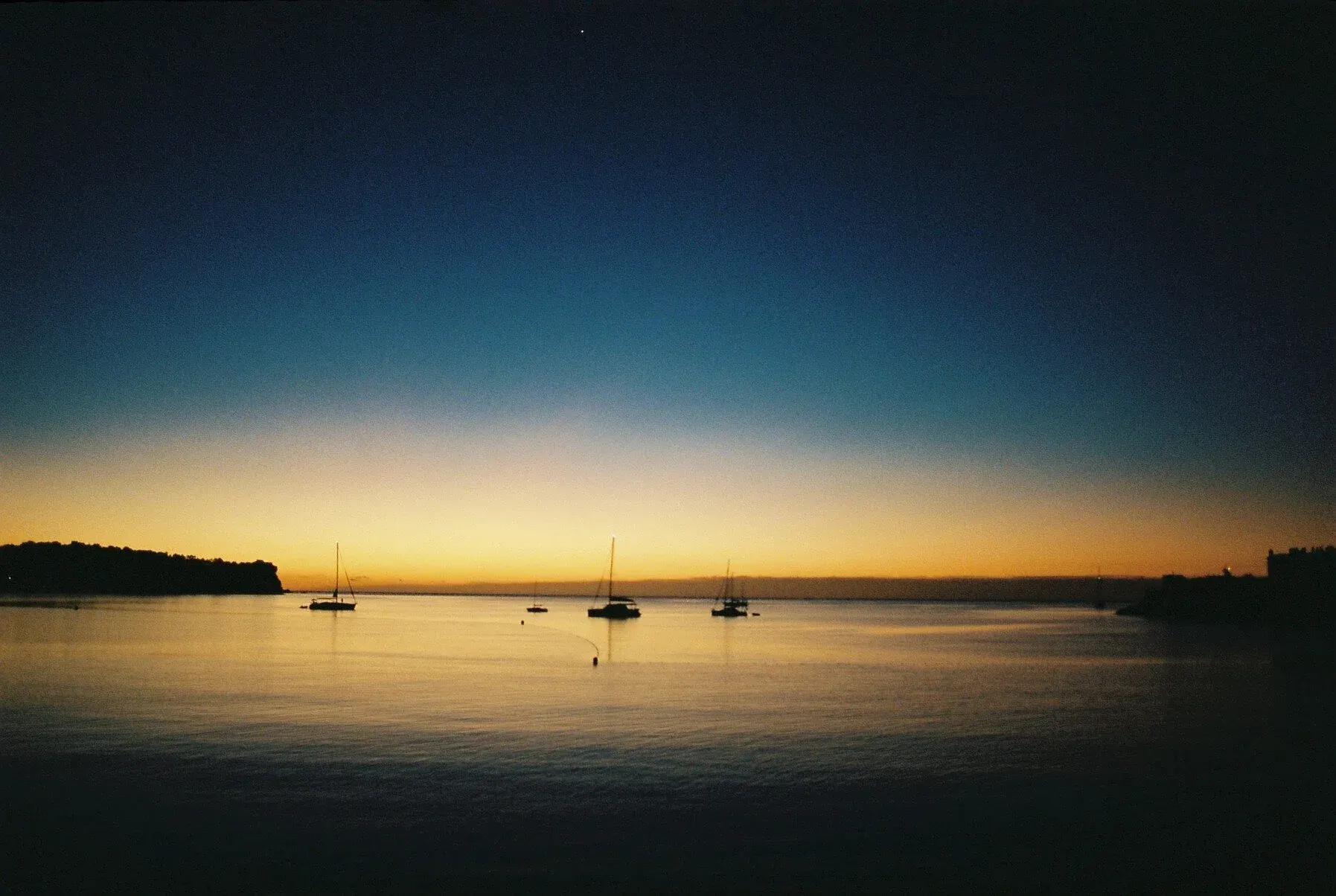 Coucher de soleil sur une mer calme avec plusieurs bateaux à voile en silhouette et un horizon lointain, ciel dégradé du jaune à l'azur.