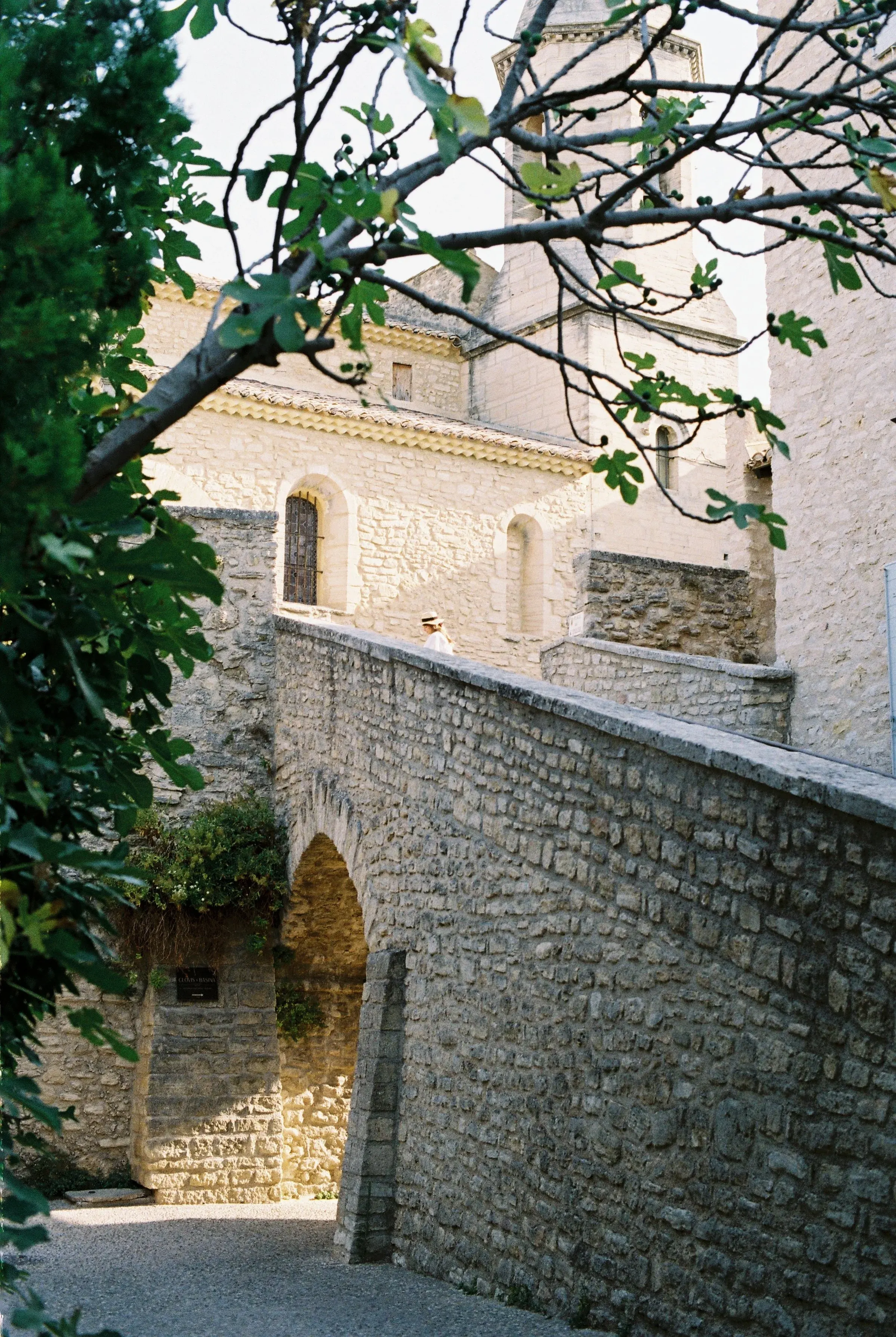 Vue de vieux bâtiments en pierre avec une arche, une petite fenêtre et un clocher en arrière-plan, entourés d'arbres et de végétation.