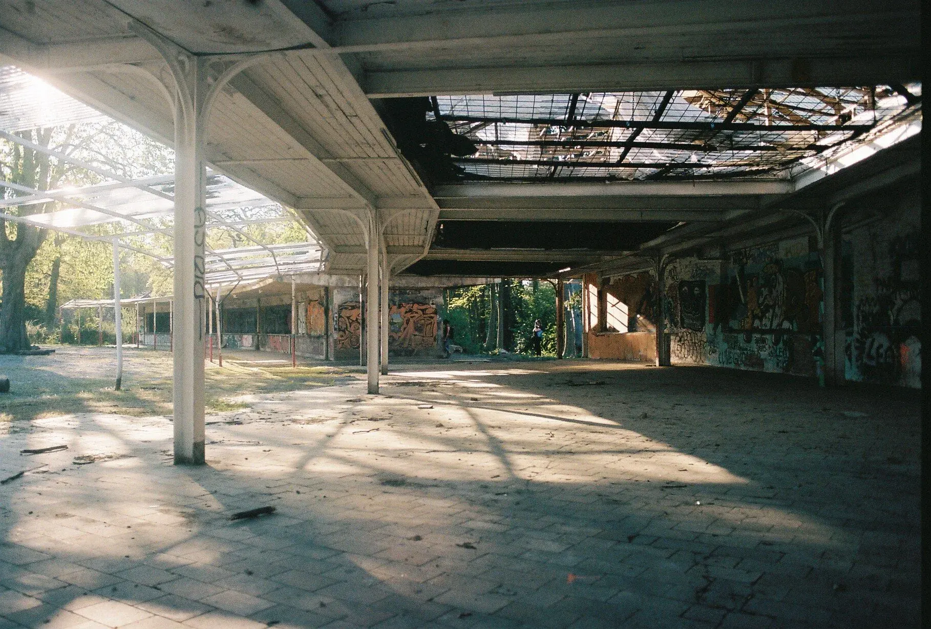Un bâtiment abandonné avec un toit partiellement effondré, recouvert de graffitis, avec des parois en béton déglinguées et un sol pavé en pierre, éclairé par la lumière du soleil filtrant à travers des arbres à l'extérieur.