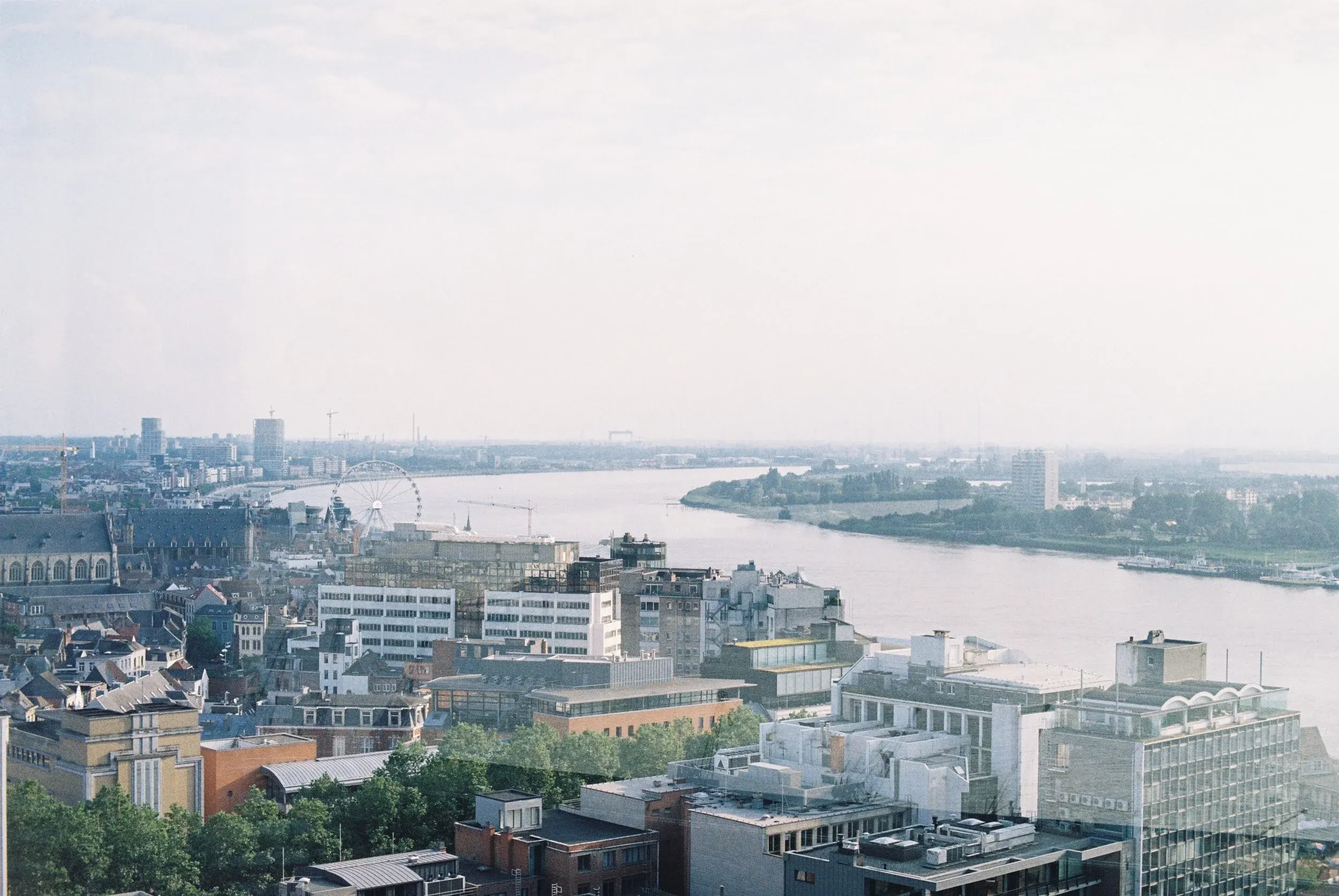 Vue aérienne d'une ville avec un fleuve, des bâtiments modernes et anciens, une grande roue et un ciel clair.