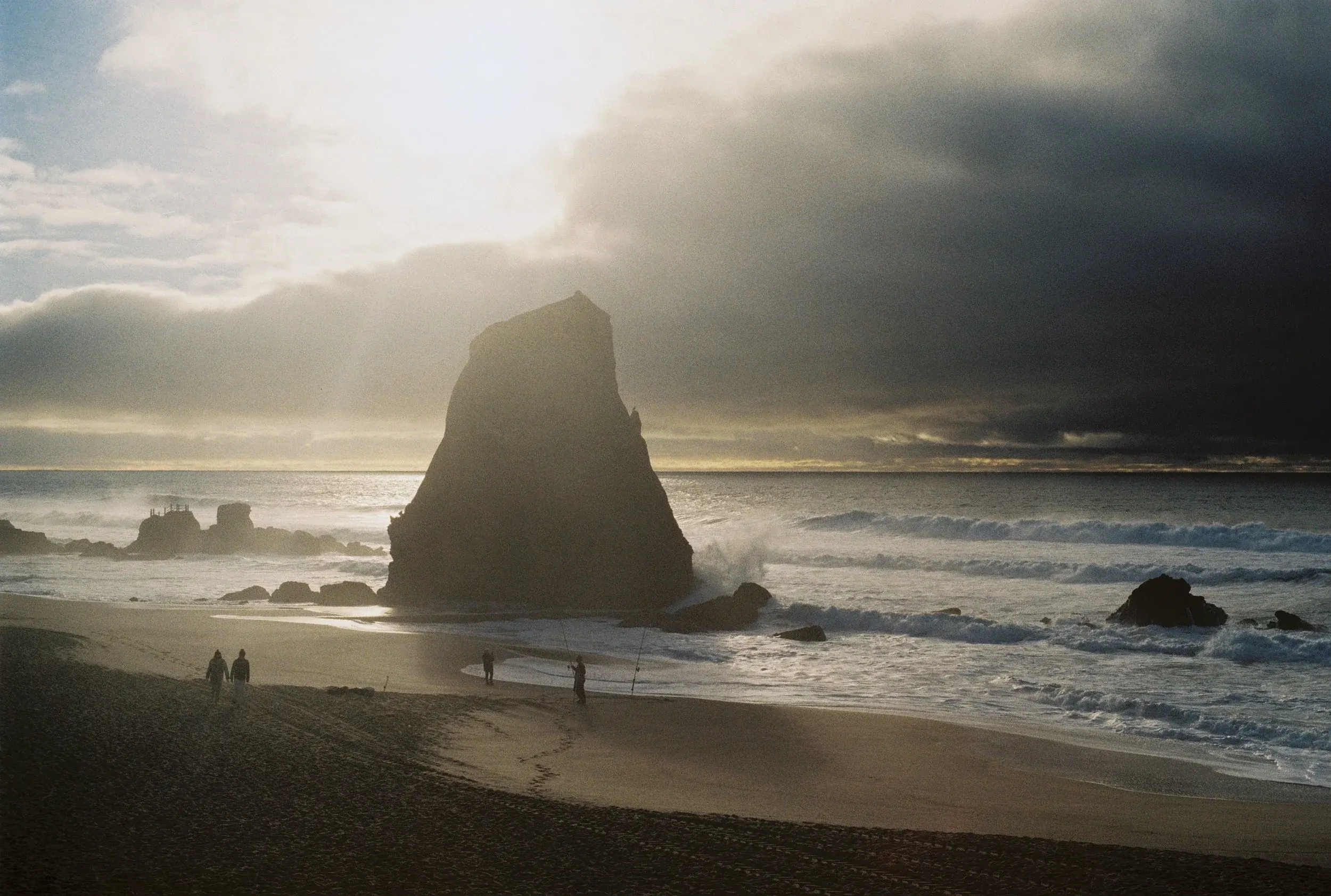 Une plage avec de grosses roches, des vagues et un ciel nuageux, quelques personnes marchant sur le sable au crépuscule.