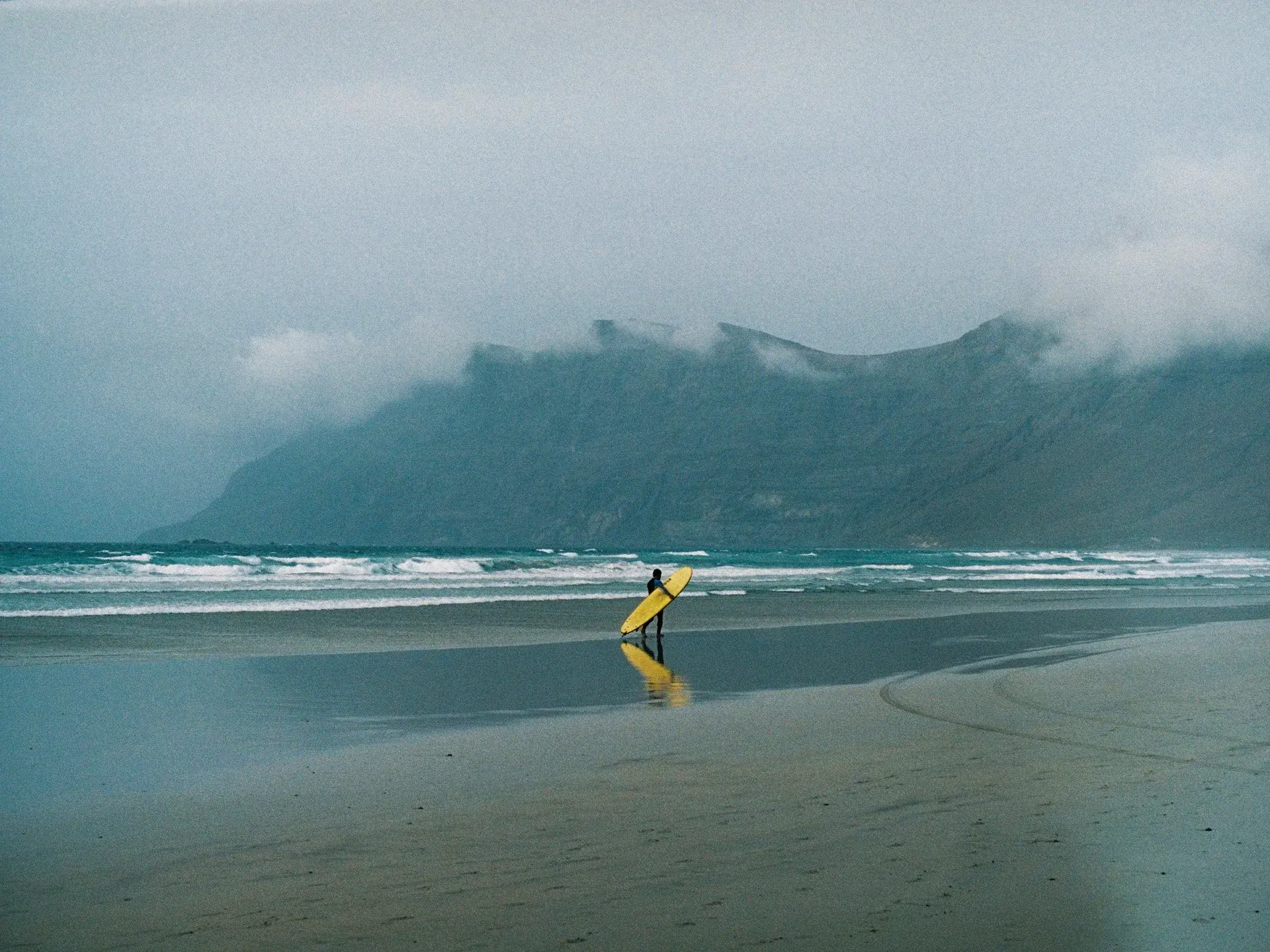 Un surfeur marchant sur la plage avec une planche de surf jaune, entouré d'une atmosphère brumeuse, avec des montagnes en arrière-plan et des vagues à l'horizon.