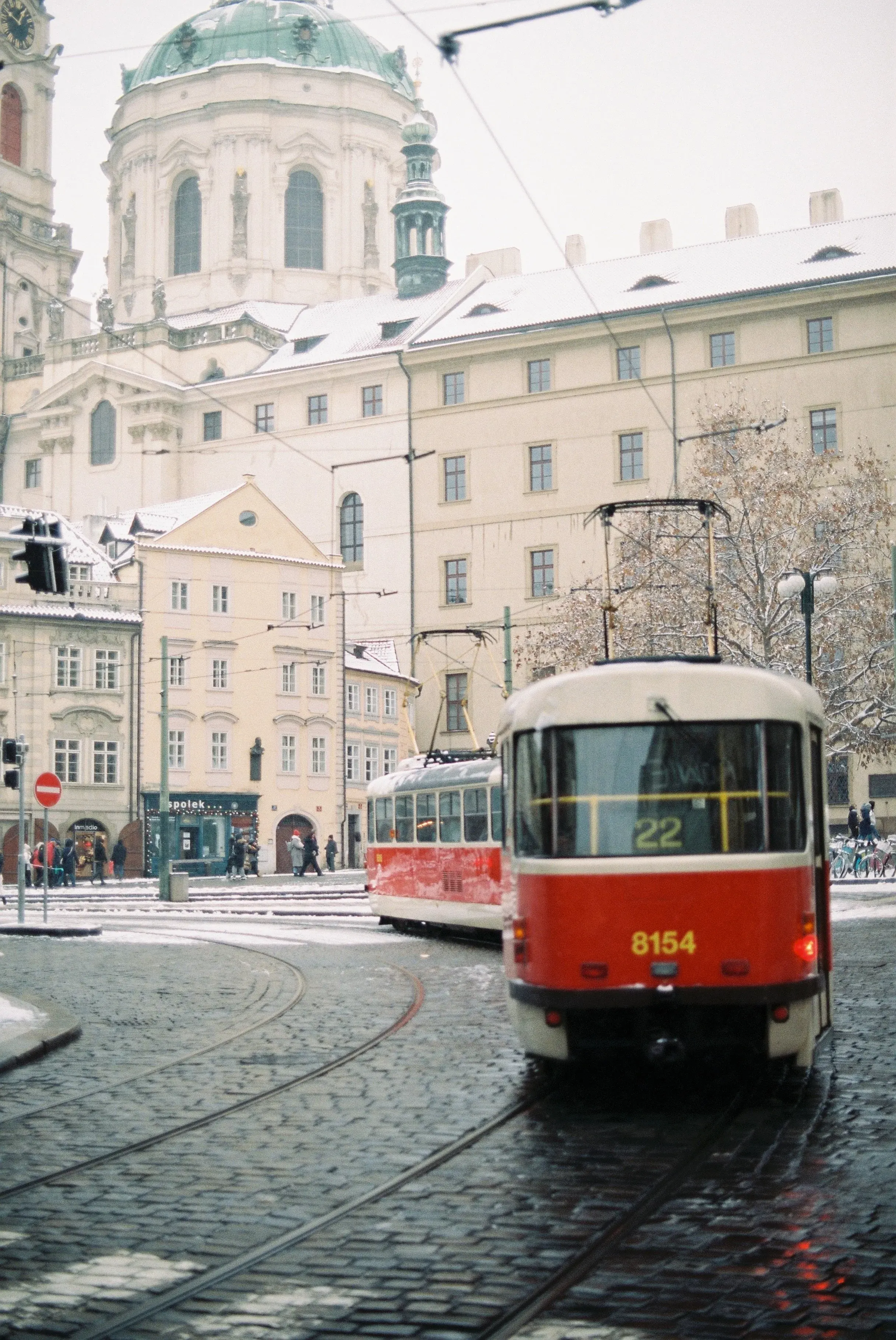 Un tram rouge et blanc circule sur des rails pavés dans une ville enneigée avec des bâtiments anciens en arrière-plan.