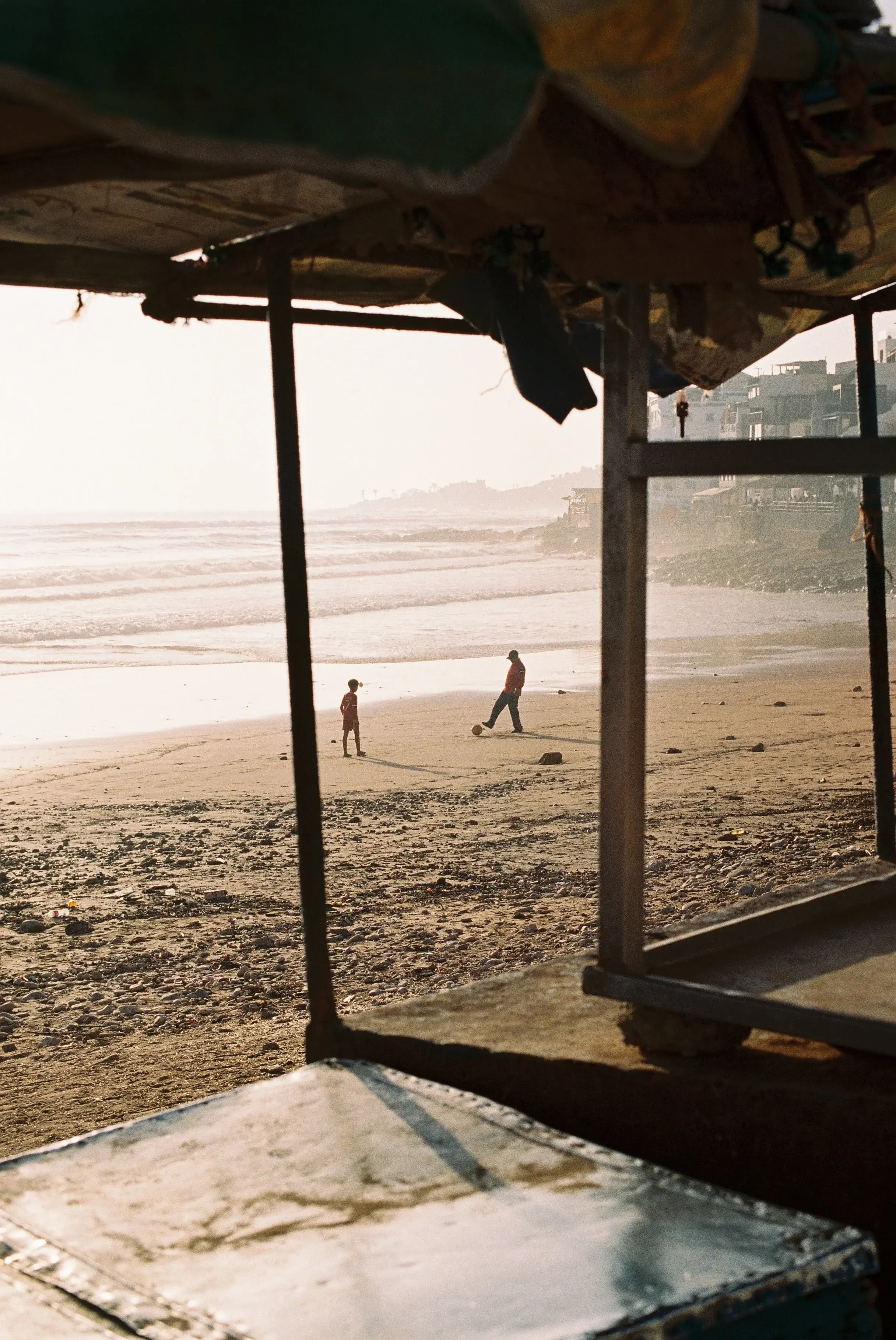 Deux enfants jouent au football sur la plage au coucher du soleil, vus à travers une structure en bois et en tissu.