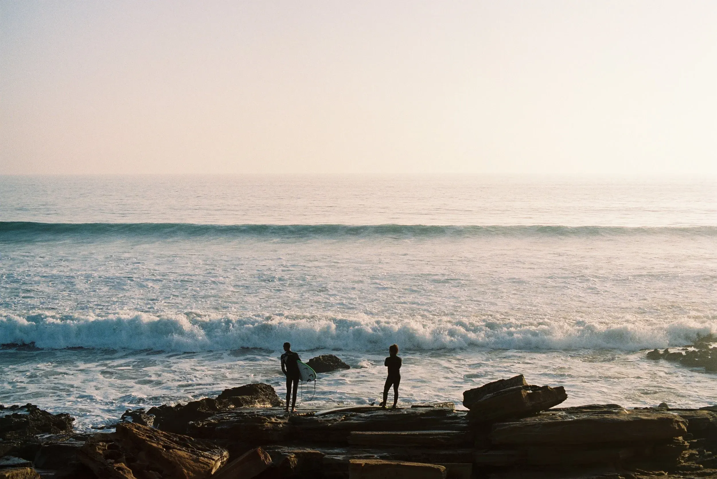 Deux personnes se tiennent sur des rochers au bord de la mer, l'une d'elles avec une planche de surf, au coucher du soleil.