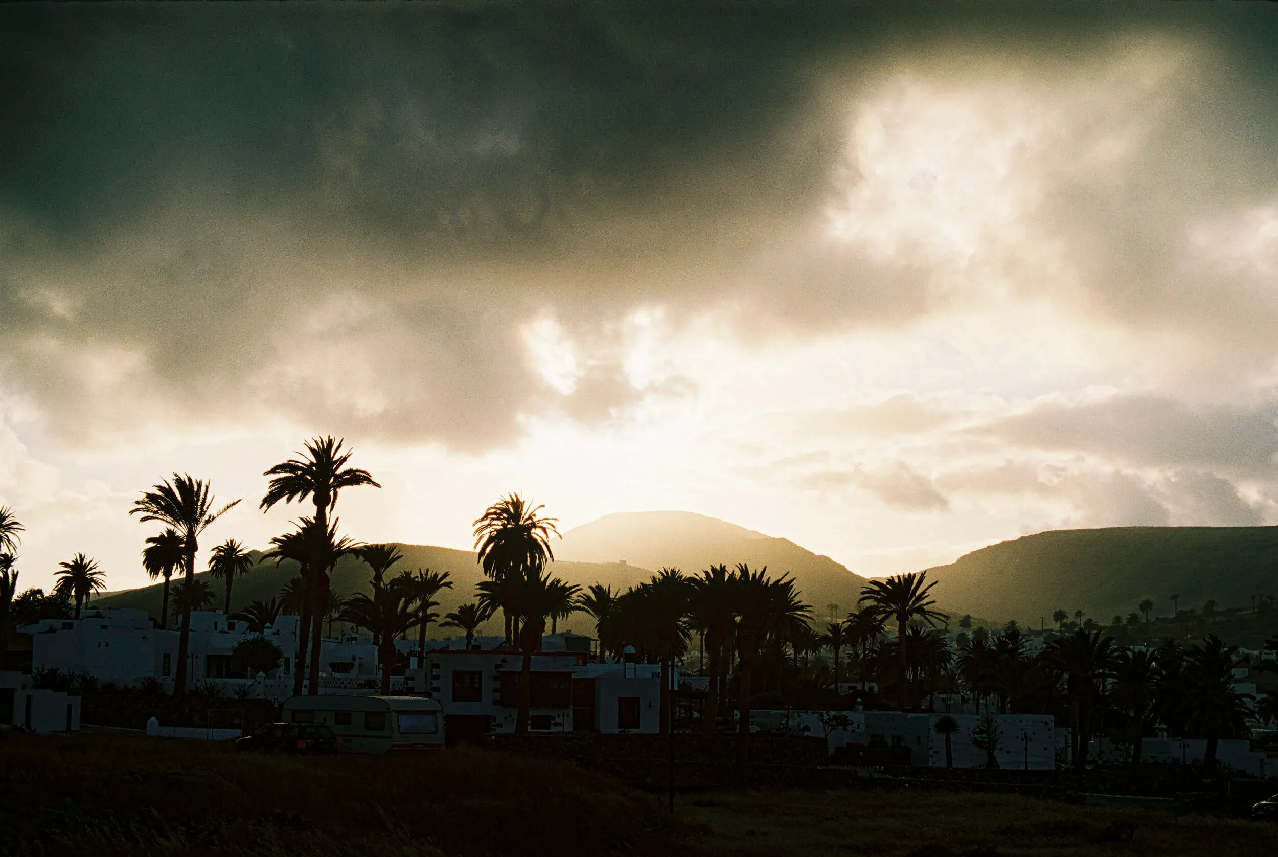 Paysage avec des palmiers, des maisons blanches et des montagnes sous un ciel nuageux au coucher du soleil.
