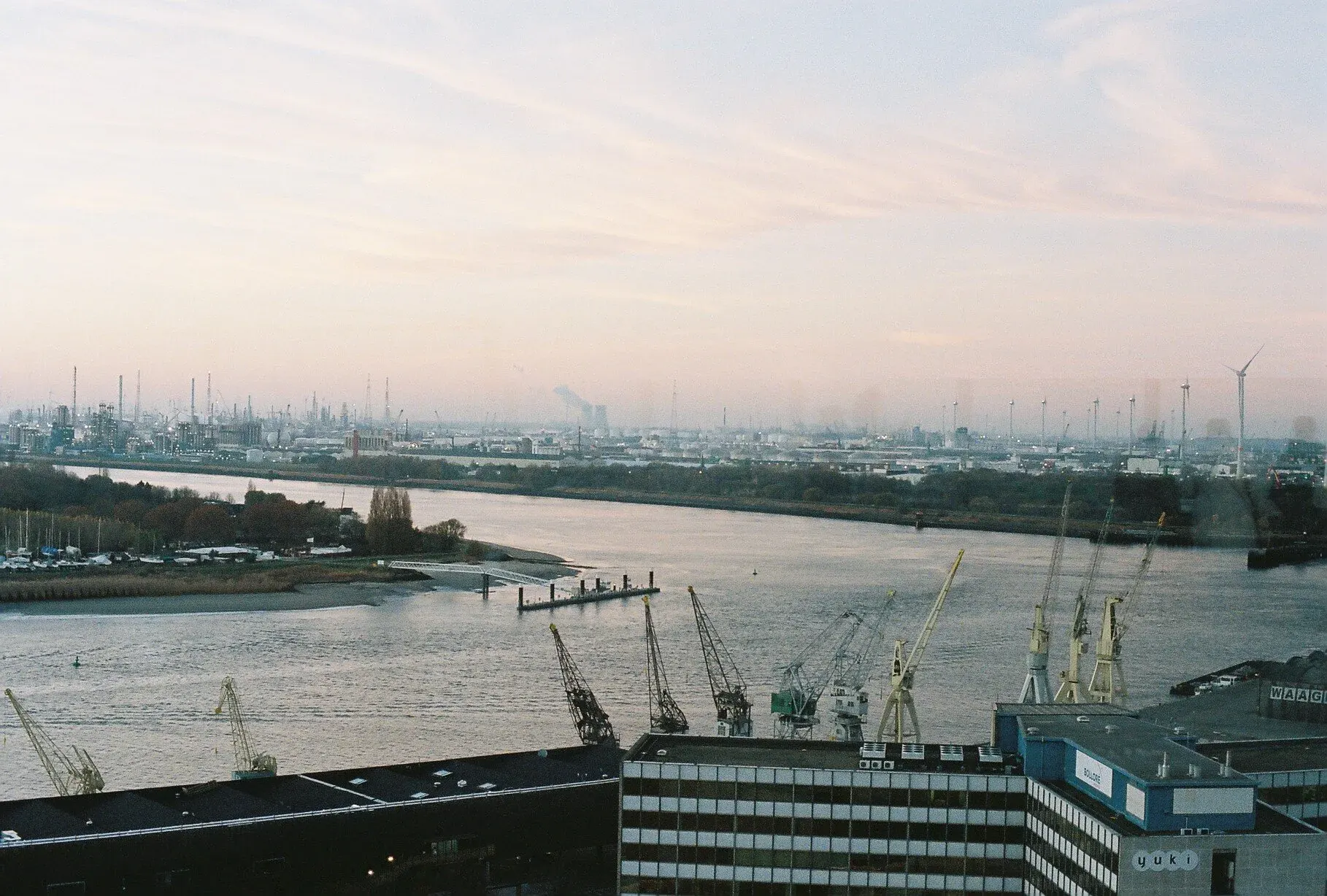 Vue d'une rivière avec une ville industrielle en arrière-plan, comprenant plusieurs grues de chantier le long du quai, des bâtiments modernes au premier plan, et un ciel légèrement nuageux.