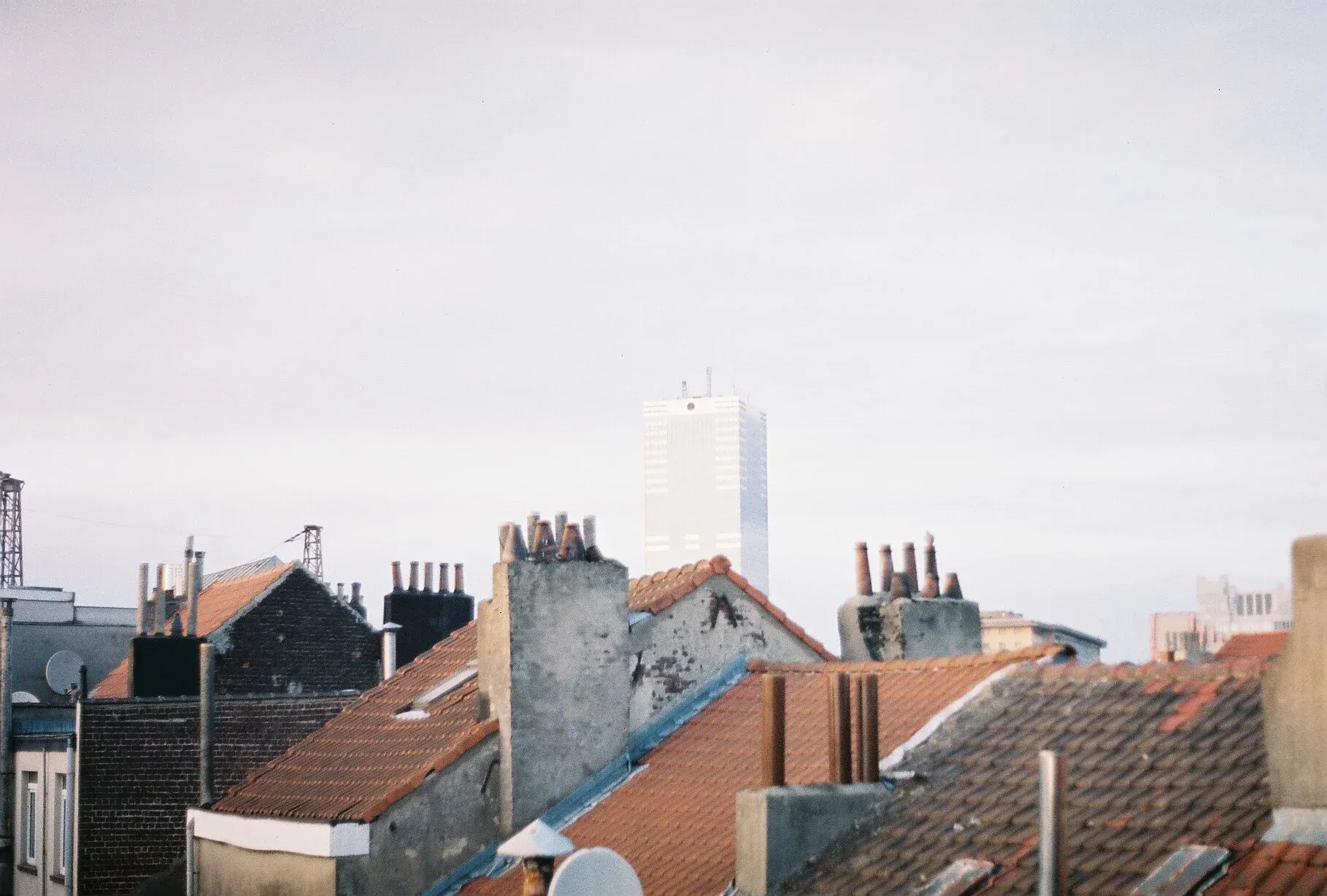 Vieux toits en tuiles avec cheminées en arrière-plan, et un grand immeuble moderne dans le fond sous un ciel nuageux.