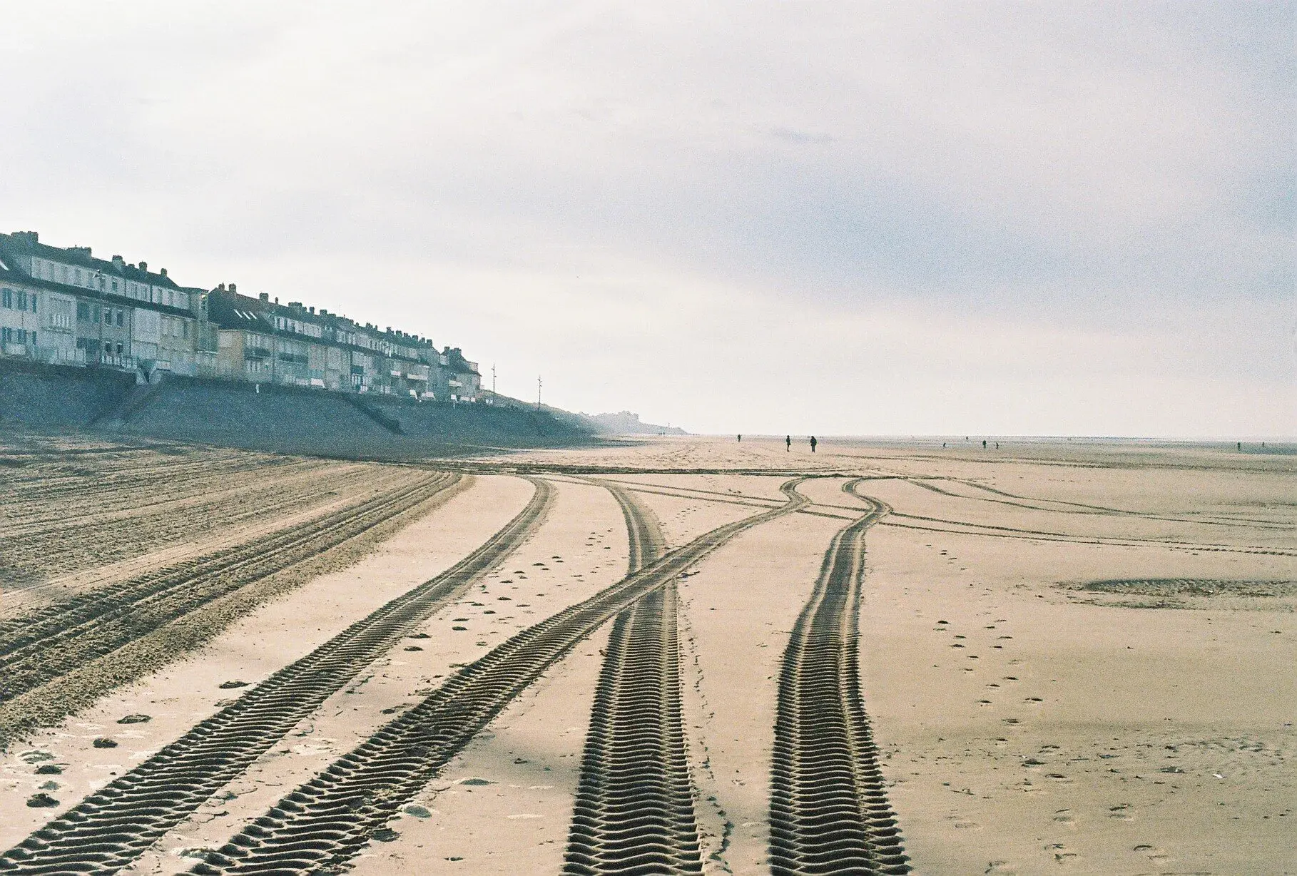 Plage avec des traces de pneus dans le sable, quelques personnes en distance et un lotissement en hauteur à gauche sous un ciel nuageux.