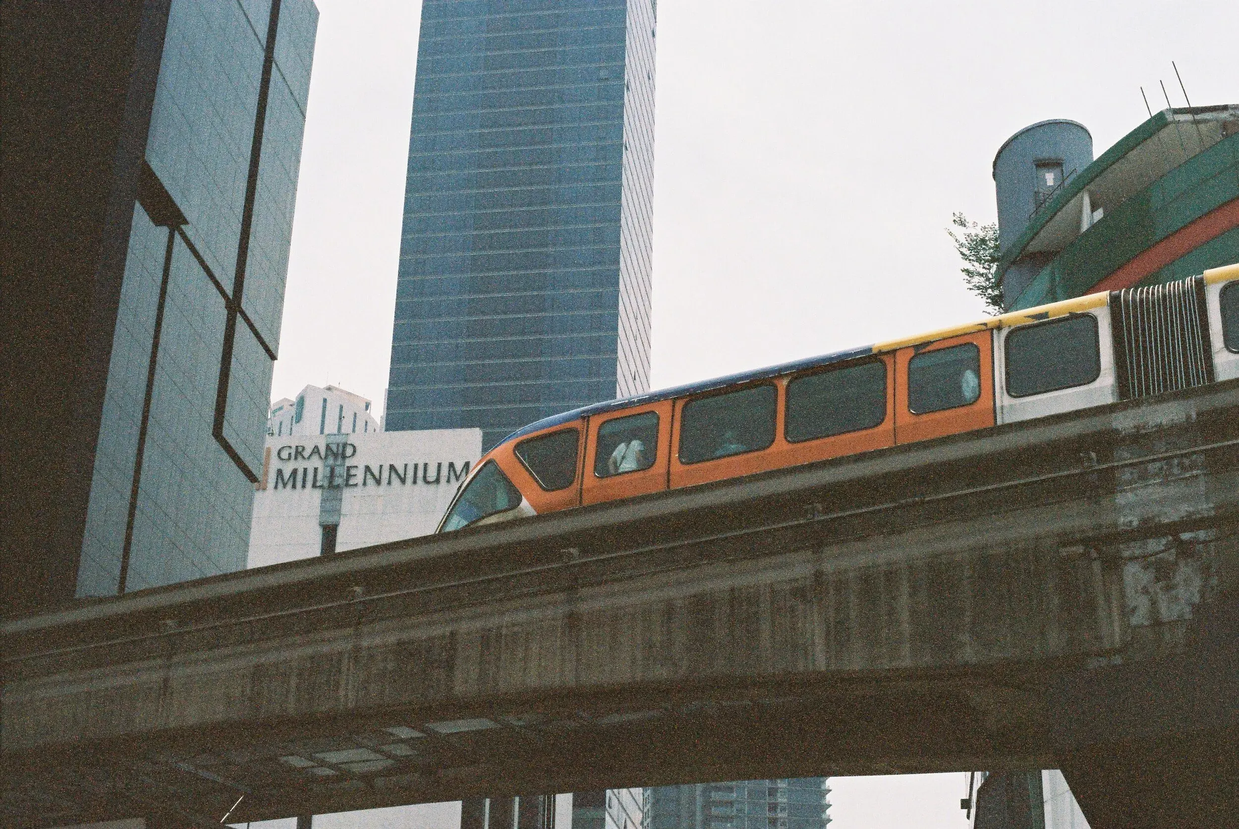 Un métro orange passant sur une ligne suspendue dans une zone urbaine avec des gratte-ciel, dont un avec un panneau indiquant 'Grand Millennium'.