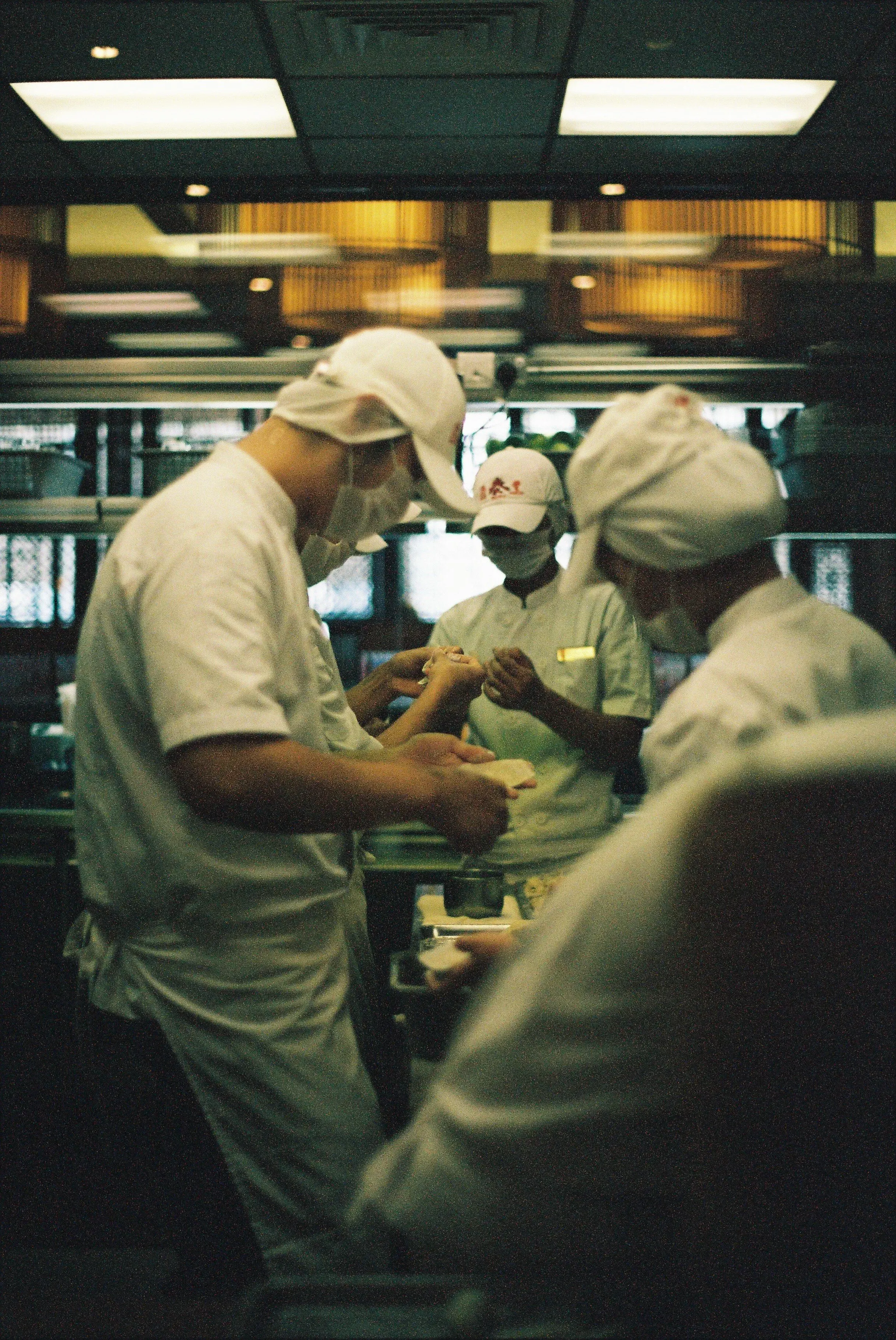 Des chef(fe)s cuisiniers en uniforme blanc, portant des masques et des chapeaux, travaillent dans une cuisine professionnelle.