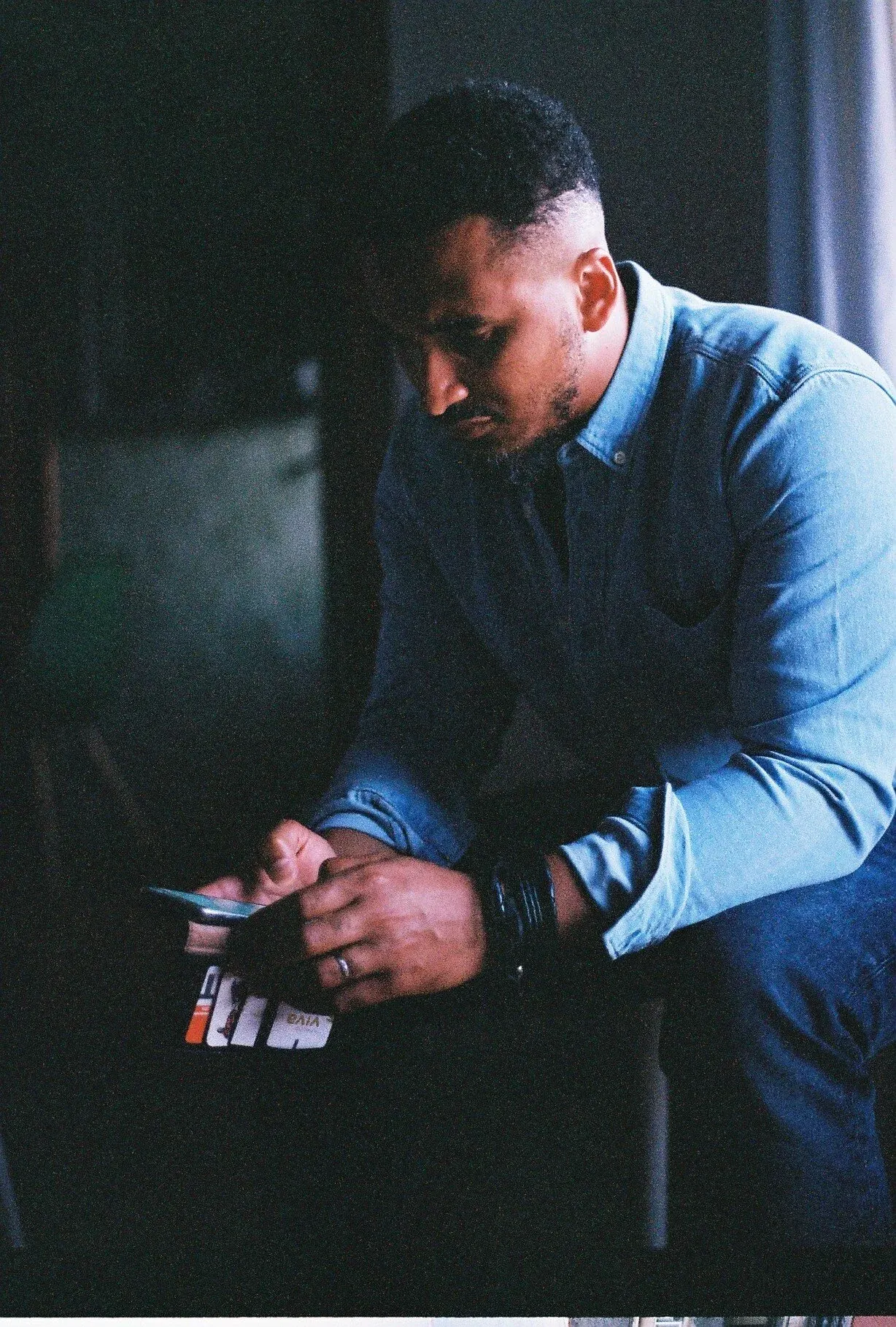 Homme assis, concentré sur son téléphone, portant une veste en jean, dans une pièce sombre.