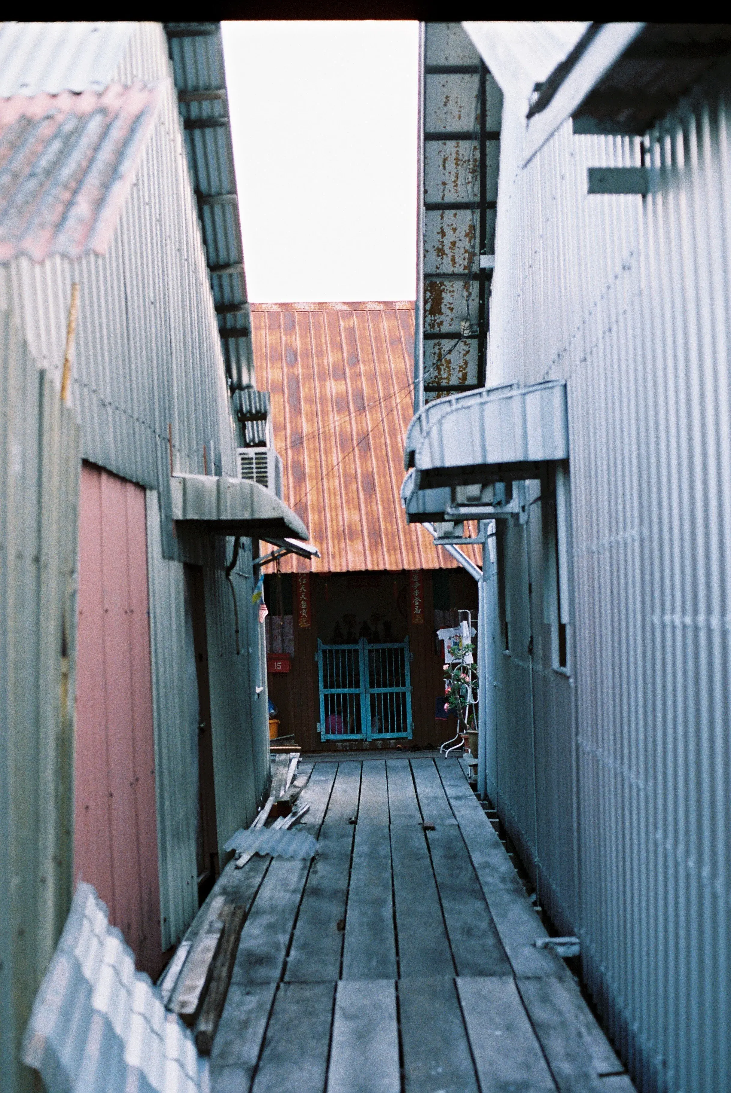 Vue d'une allée étroite bordée de bâtiments en tôle métallique avec un chemin en bois menant à une porte bleue dans un centre chinois.