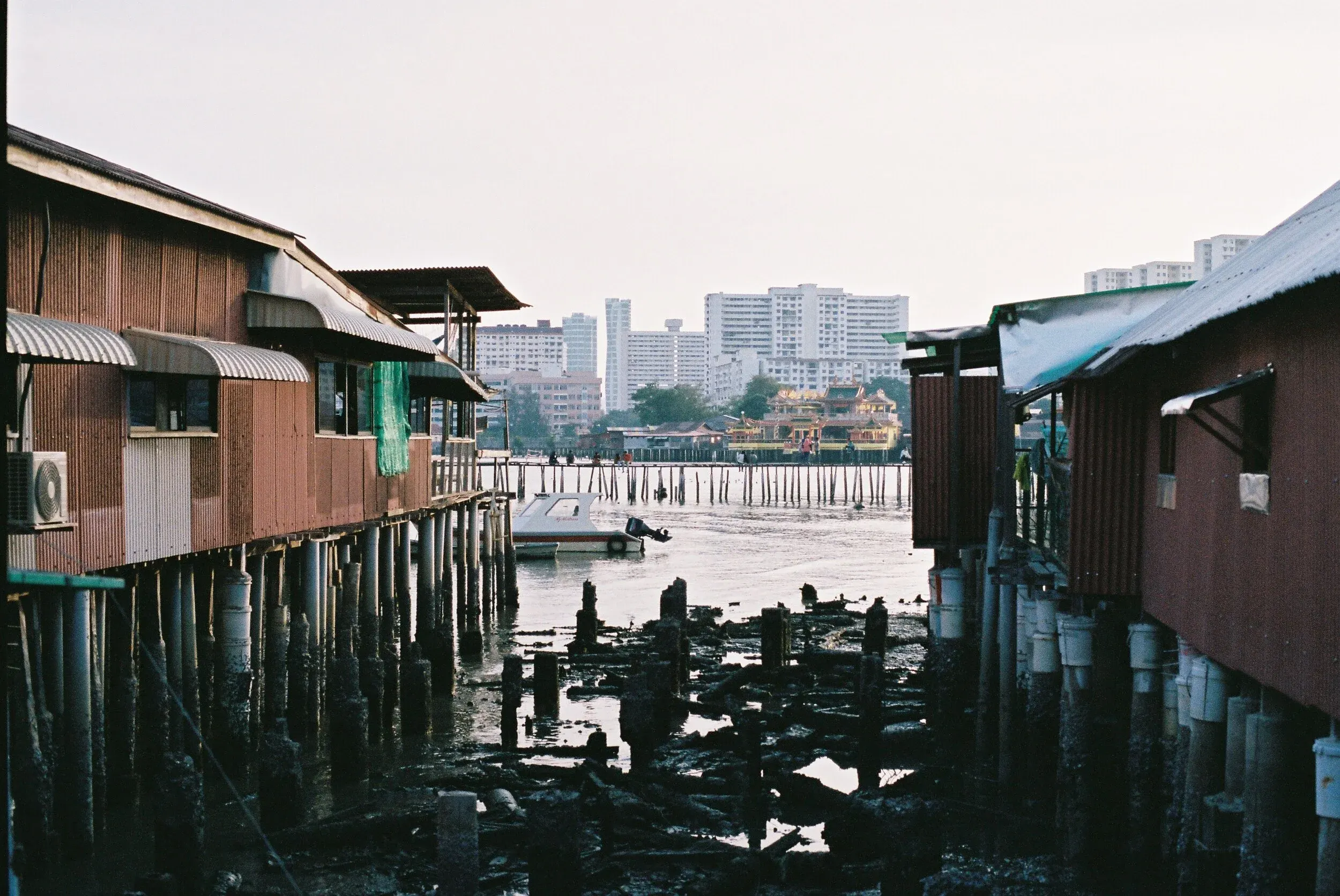 Maisons en bois sur pilotis au bord de l'eau, avec un bateau, eau sombre en dessous, et bâtiments modernes en arrière-plan.