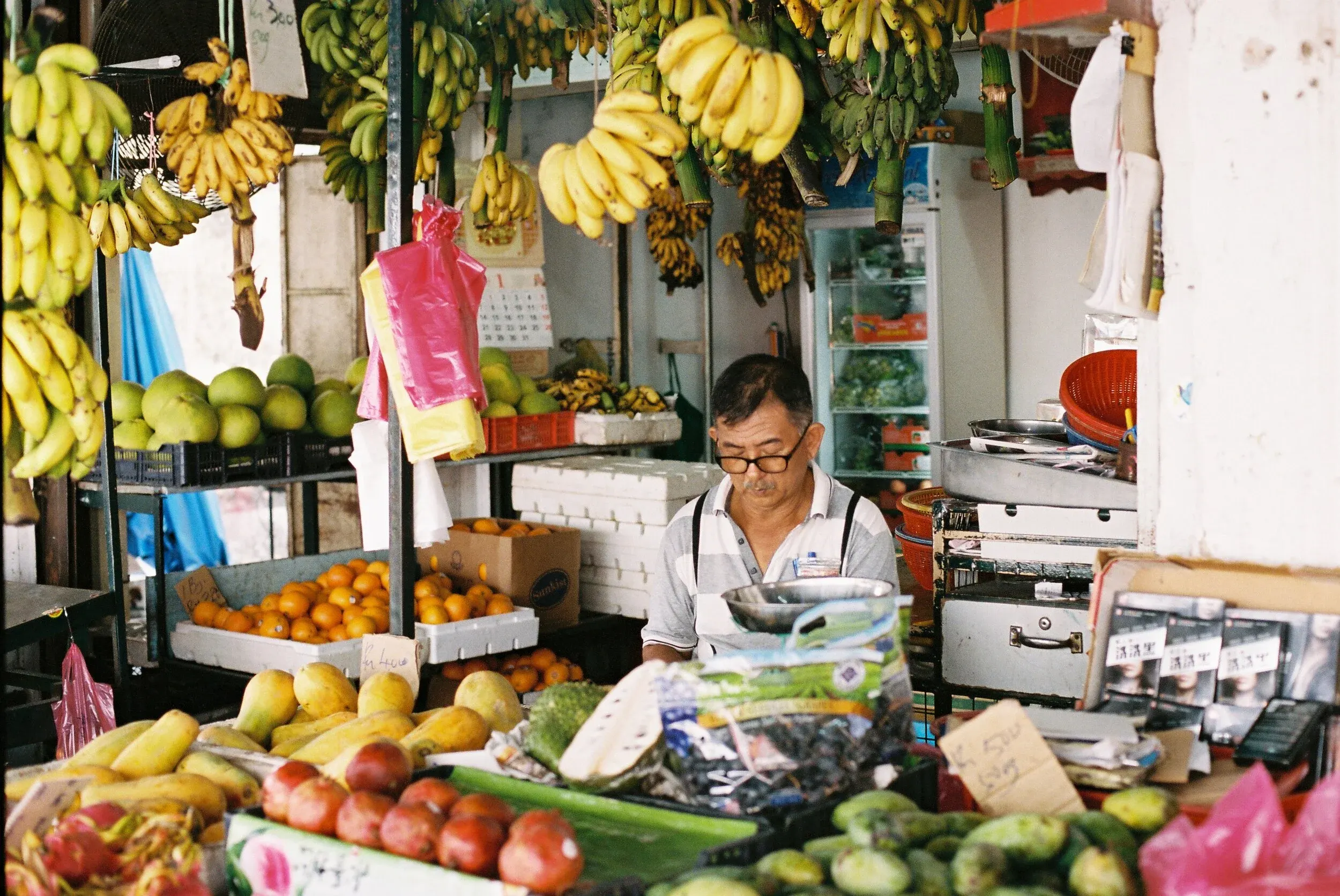 Marché de fruits avec des bananes, oranges, fruits exotiques et un vendeur âgé portant des lunettes.