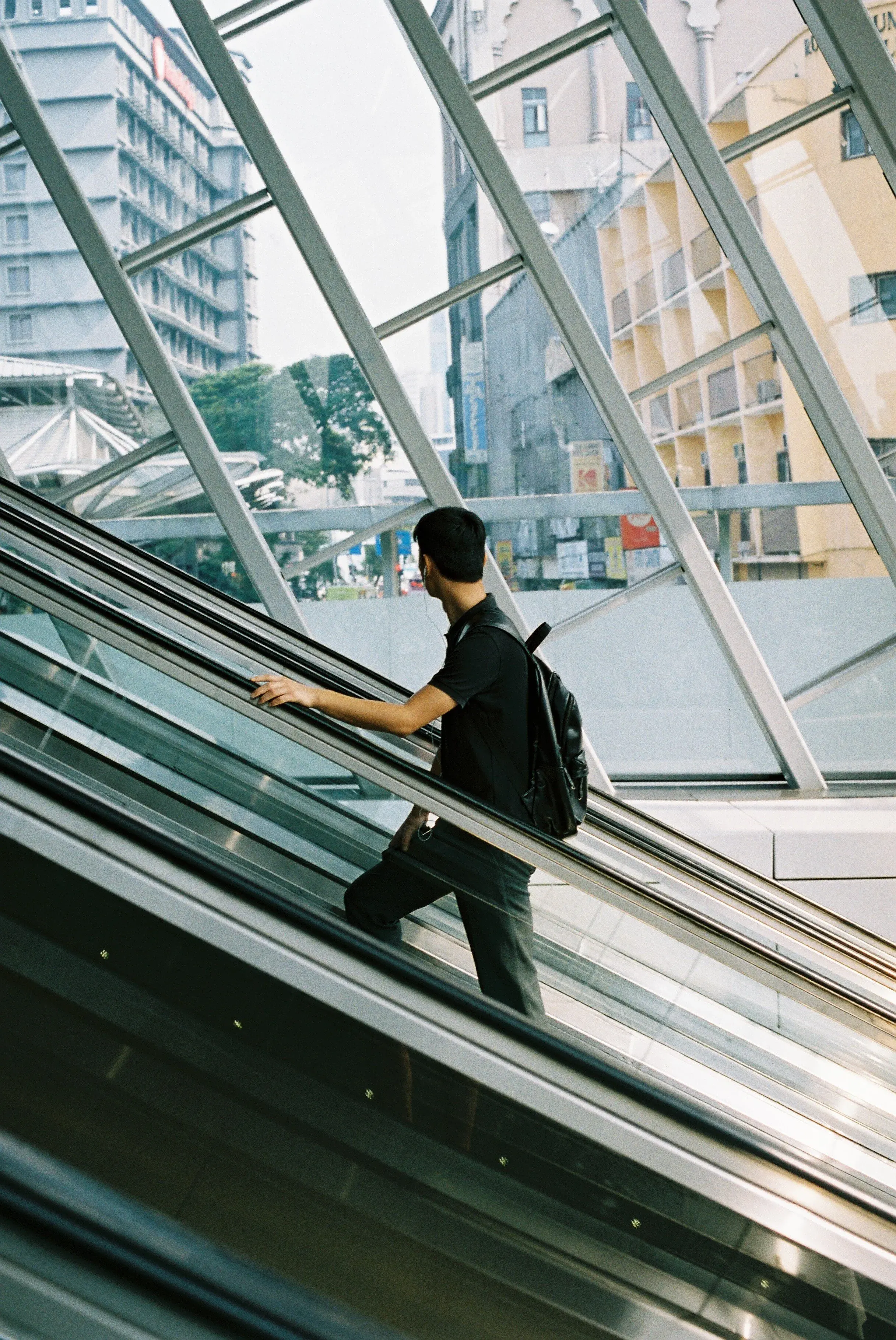 Un jeune homme avec un sac à dos descend un escalator dans un bâtiment moderne avec des murs en verre et des structures métalliques.