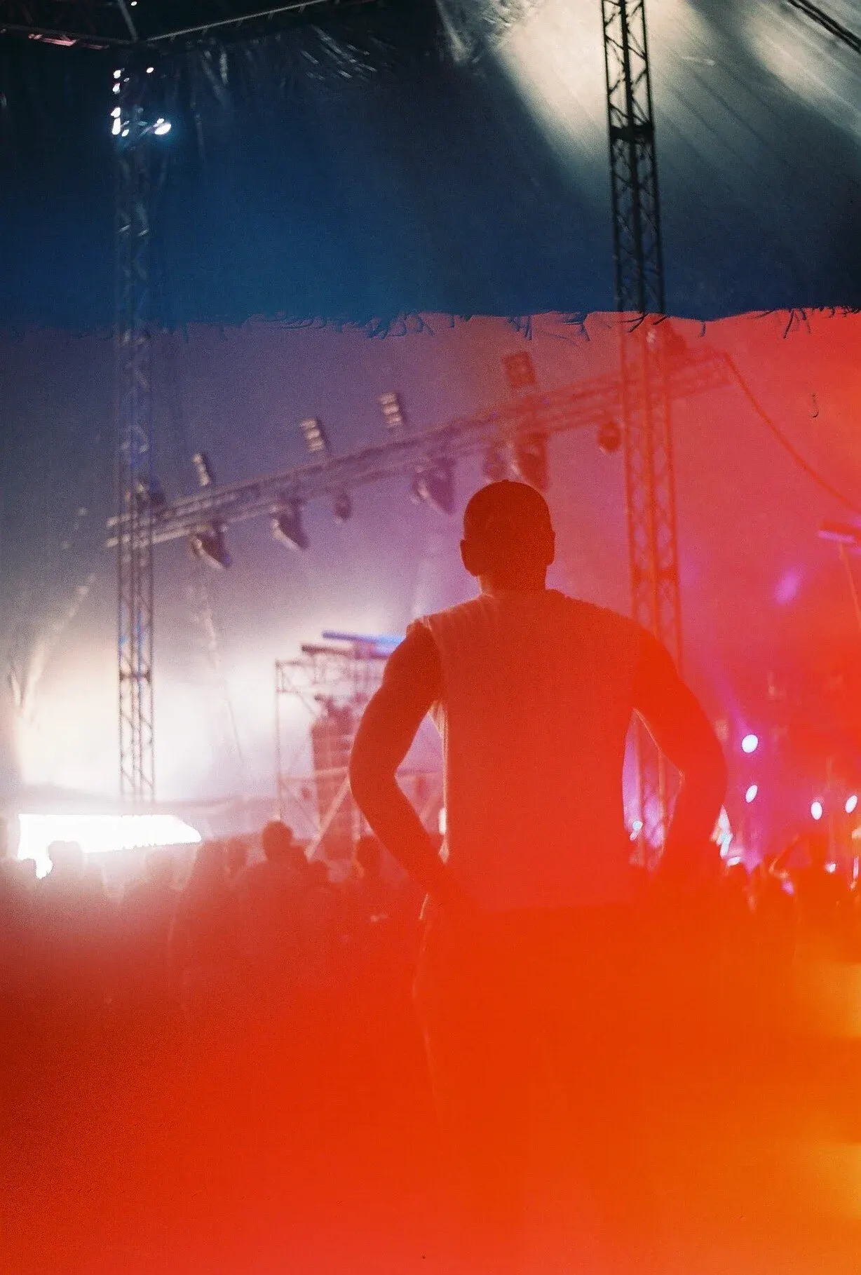 Silhouette d'une personne debout dans une scène de concert ou de spectacle avec des lumières colorées et une structure métallique en arrière-plan.