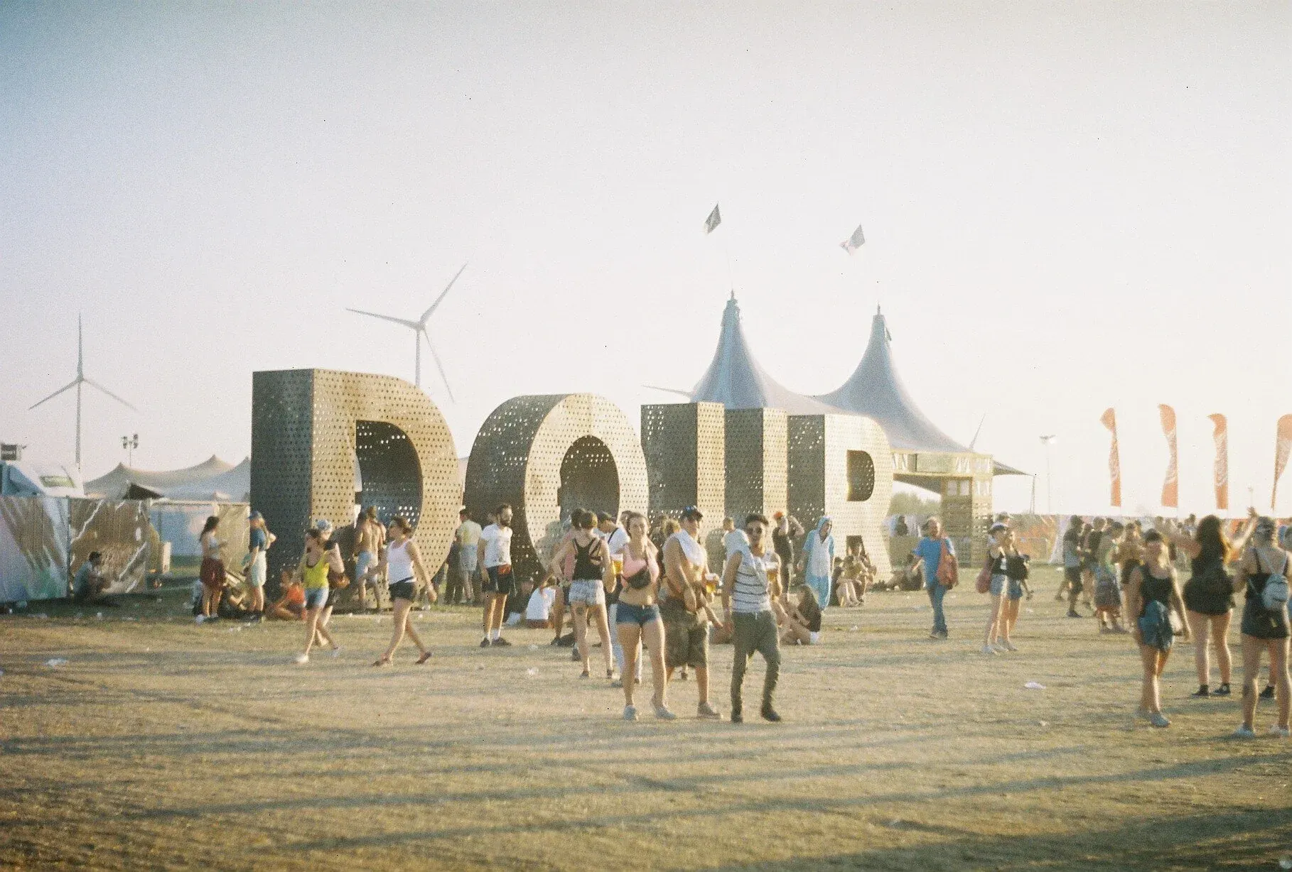 Festival en plein air avec grande structure en forme de lettres DOUB sous un ciel clair, de nombreuses personnes en tenue légère, tentes, drapeaux et moulins à vent en arrière-plan.