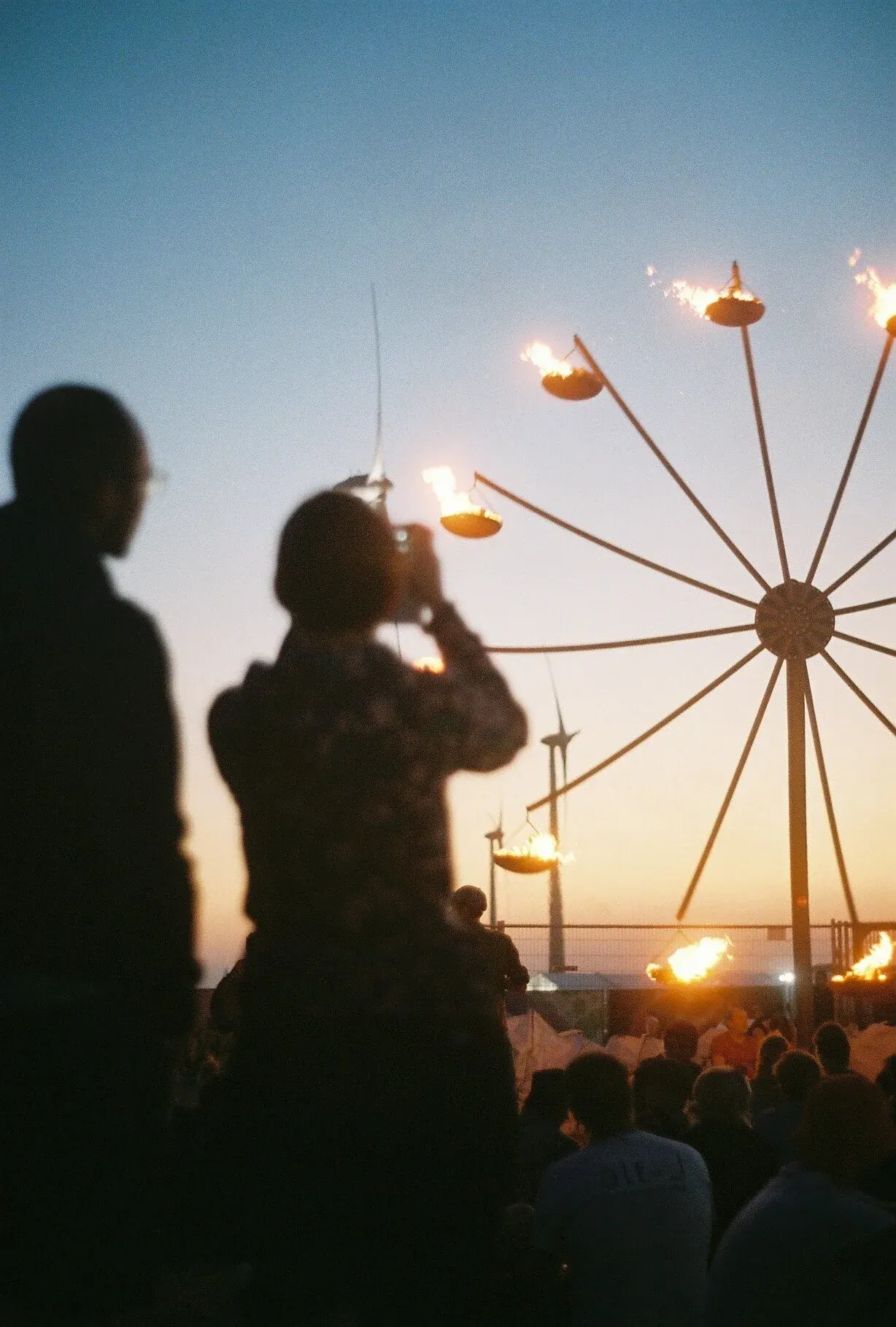 Groupe de personnes regardant une fête foraine avec des manèges en fête et des torches enflammées lors du crépuscule.