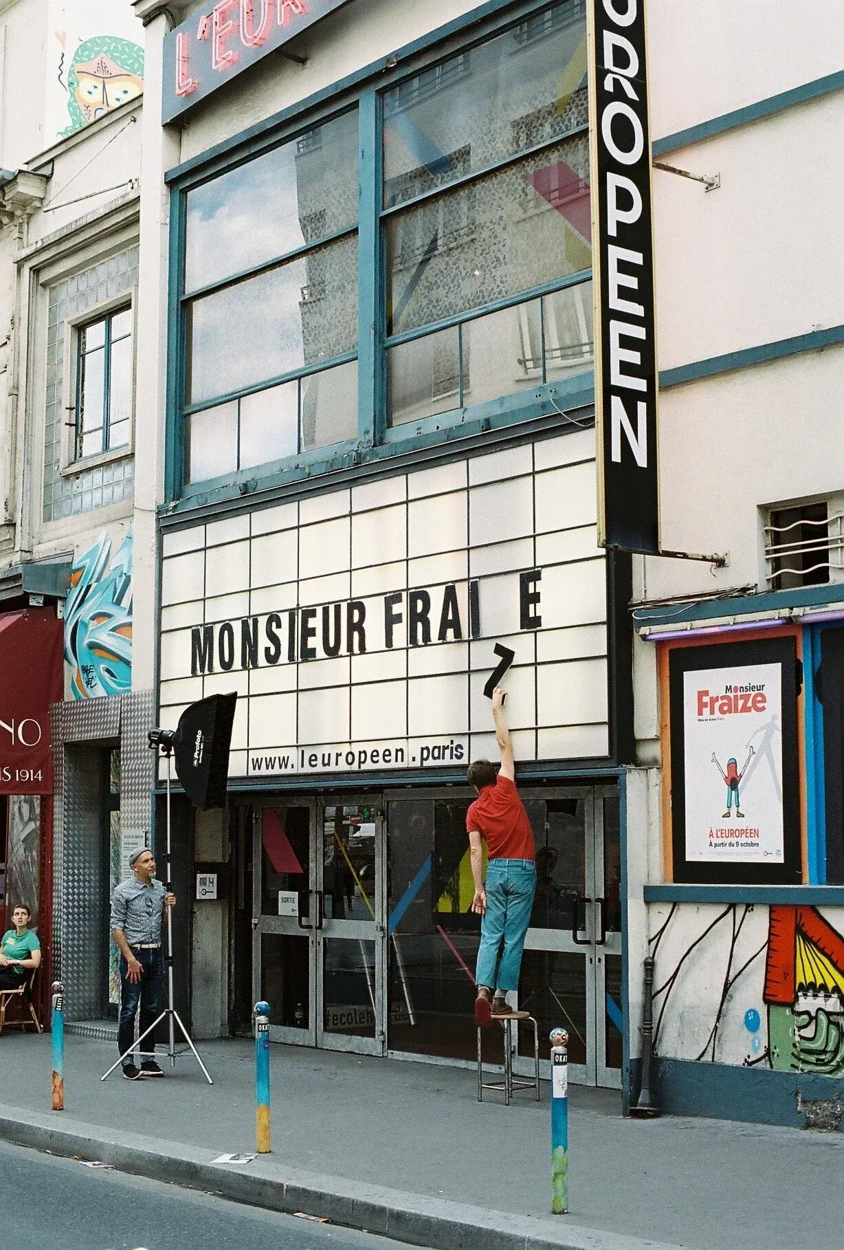 Une scène urbaine devant un bâtiment à Paris, avec un homme en tenue rouge et pantalon bleu clair, utilisant un escabeau pour coller une lettre manquante sur un panneau publicitaire. Trois autres personnes sont présentes : un homme avec un manteau gr