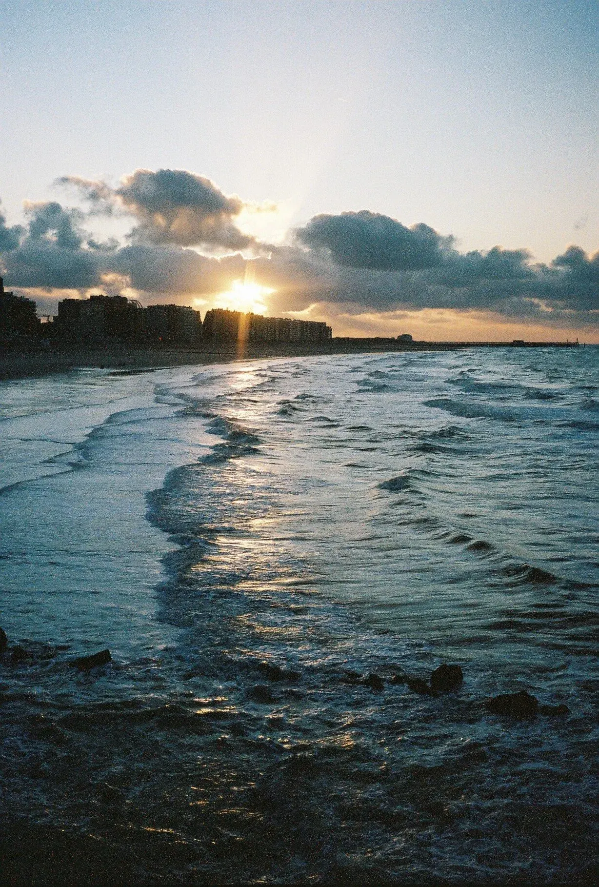 Coucher de soleil sur l'océan avec des nuages et une ligne de bâtiments à l'horizon, vagues s'écrasant sur la plage.