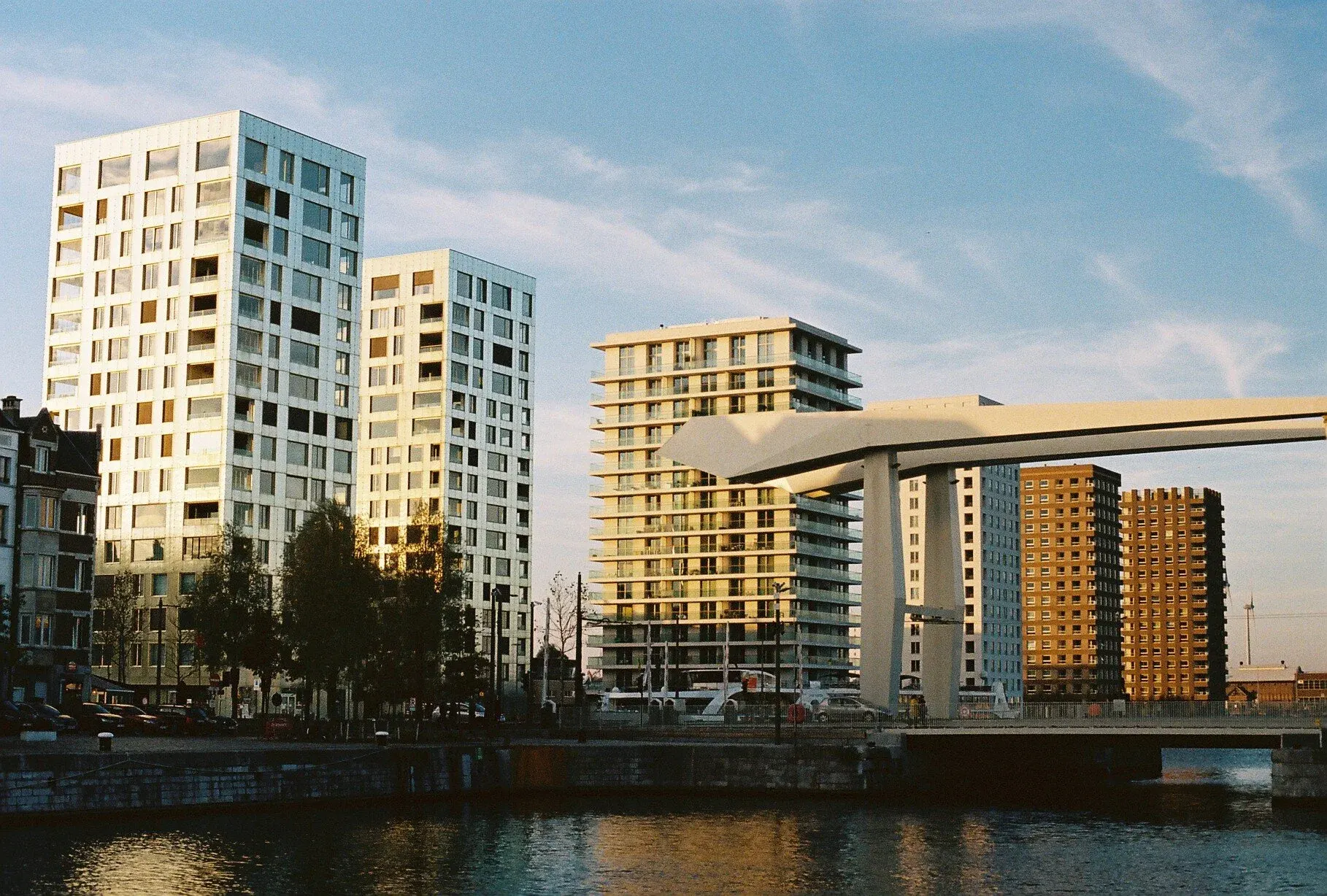Vue urbaine avec plusieurs bâtiments résidentiels modernes le long d'une rivière, en fin d'après-midi, avec un ciel bleu et quelques nuages.