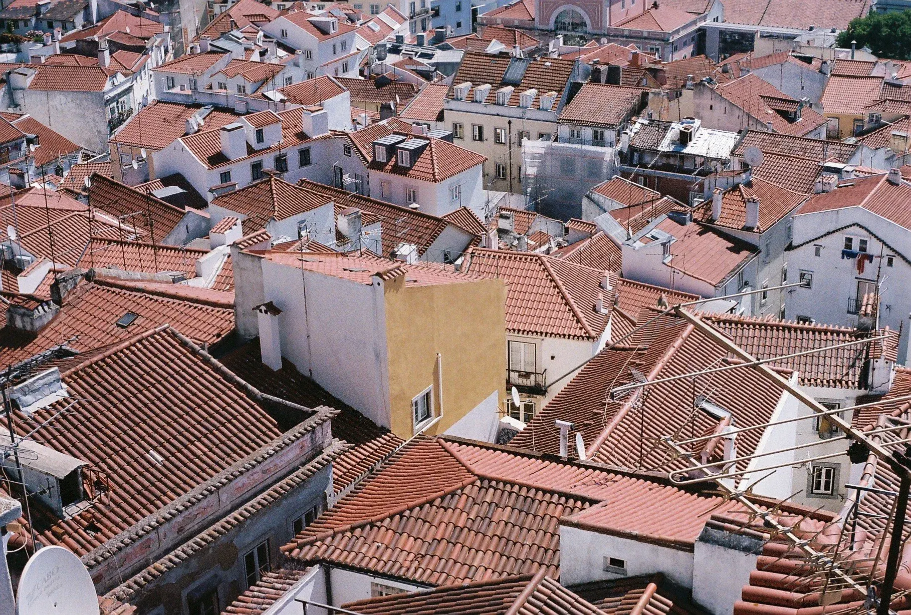 Vue aérienne d'un quartier résidentiel avec de nombreux toits en tuiles orange, quelques bâtiments avec des murs blancs et un bâtiment en jaune. On voit aussi plusieurs antennes et fenêtres.