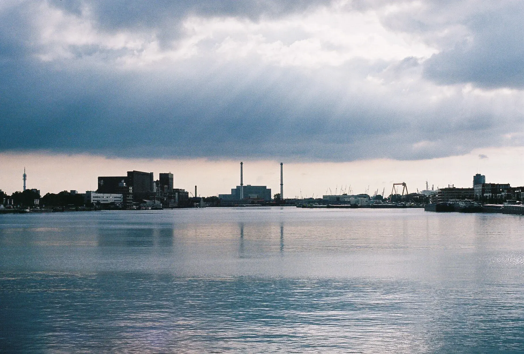 Une ville industrielle avec des usines, des bâtiments et un port, vue depuis une rivière ou un lac par un jour nuageux.