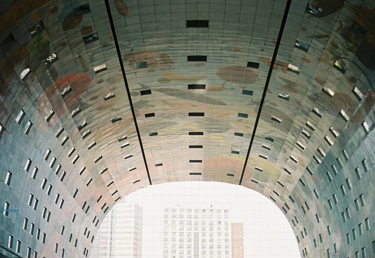 Vue depuis l'intérieur d’un tunnel au plafond métallique ondulé, avec de petites fenêtres rectangulaires et un aperçu de bâtiments en dehors.