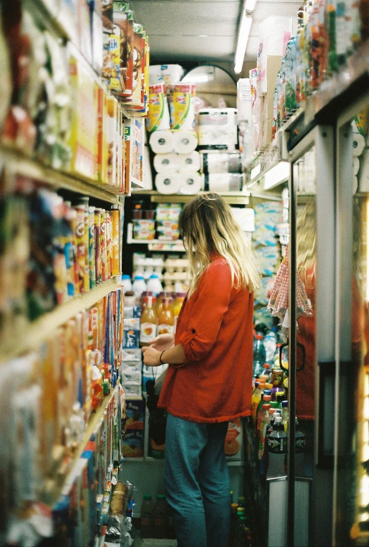 Une femme avec des cheveux longs et blonds, portant un manteau rouge et un jean bleu, regarde une bouteille dans une épicerie, entourée de rayonnages d'aliments.