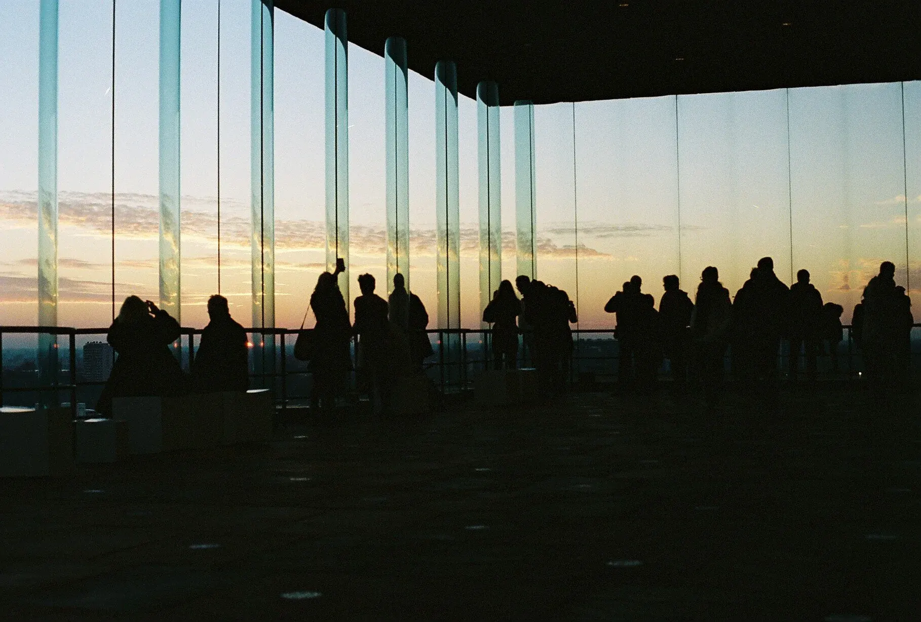 Silhouettes de personnes regardant l'horizon lors du coucher du soleil à l'intérieur d'un bâtiment moderne avec de grandes fenêtres verticales.