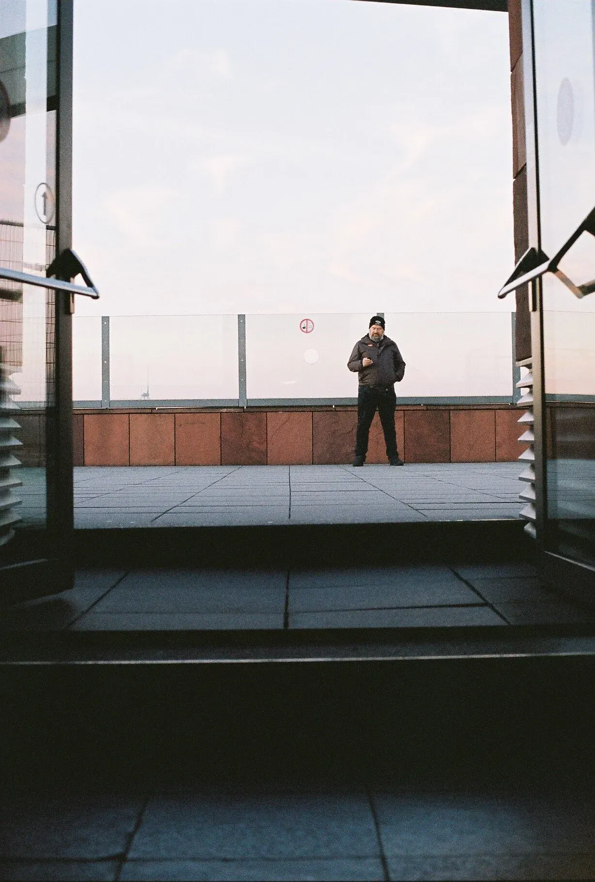 Une vue depuis l'intérieur d'un bâtiment, regardant vers un homme debout à l'extérieur sur une terrasse en journée, portant un manteau et un bonnet, avec un ciel clair en arrière-plan.