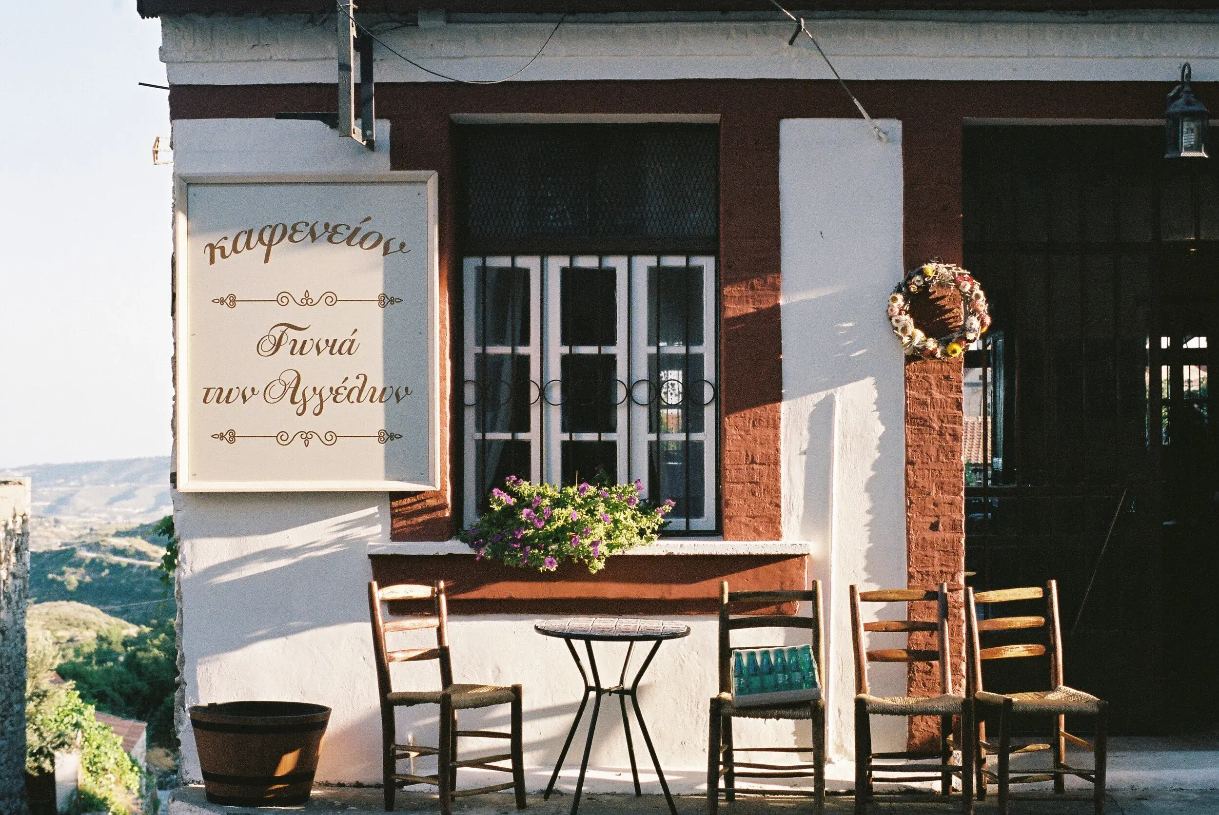 Une façade de café ou taverne avec un panneau écrit en grec, une fenêtre, une décoration florale en forme de couronne, quatre chaises en bois, une petite table, et un pot de fleurs, sous une lumière de coucher de soleil.