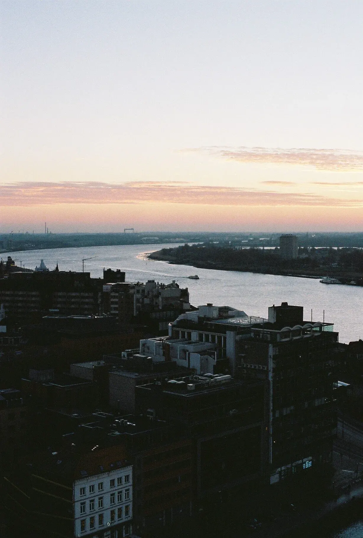 Vue d'une rivière au coucher du soleil traversant une ville avec des bâtiments modernes et des bateaux sur l'eau.